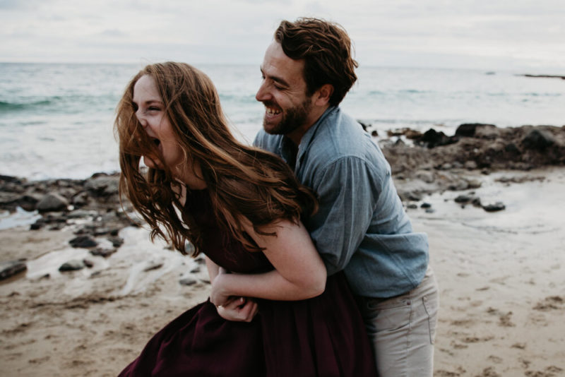 family photographer, young couple being playful on the beach, man embraces woman from behind as she laughs